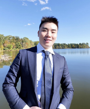 image of Weikang Zhang in front of a large blue-green lake, bordered by trees, smiling at the camera